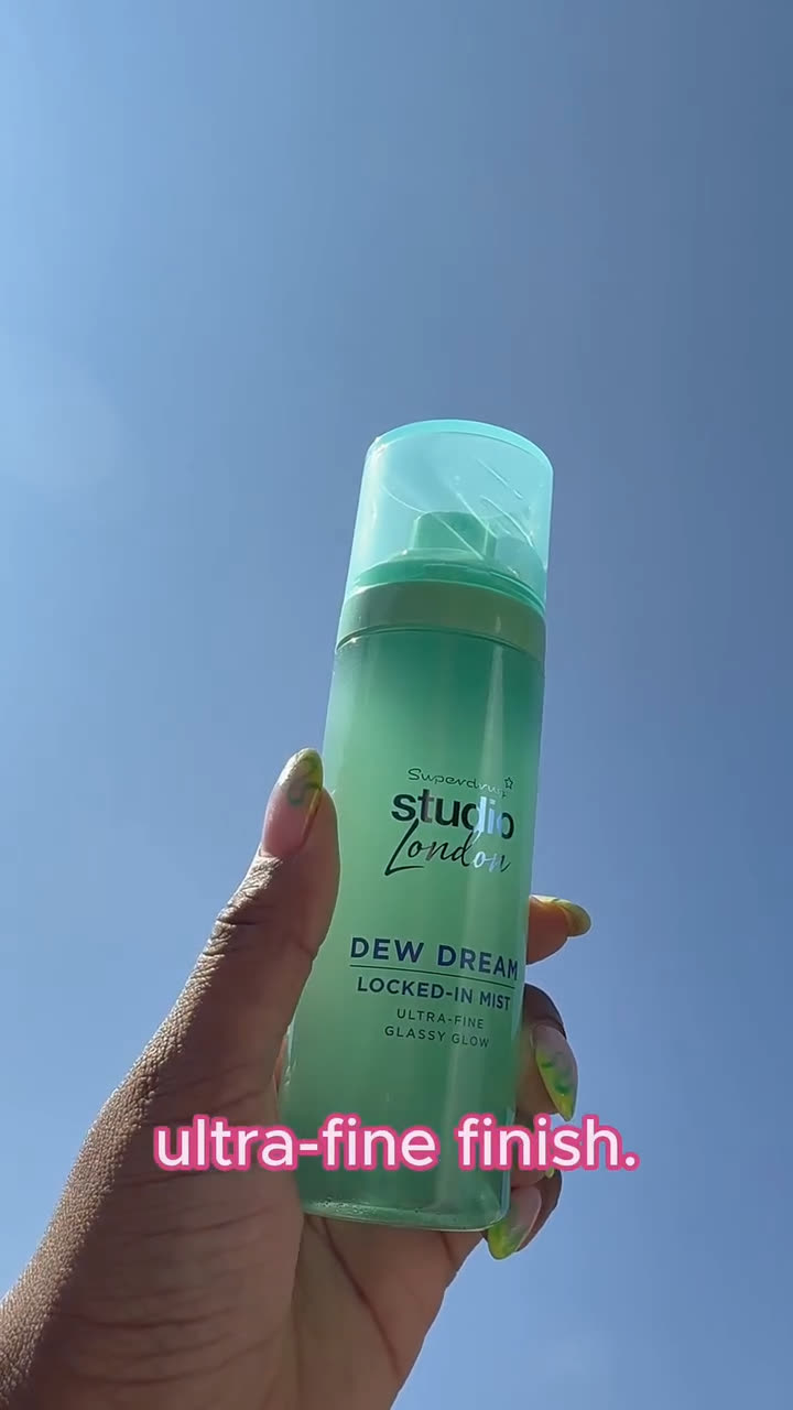 A hand holding the Studio London Dew Dream Locked-In Mist bottle against a blue sky, with the caption ultra-fine finish